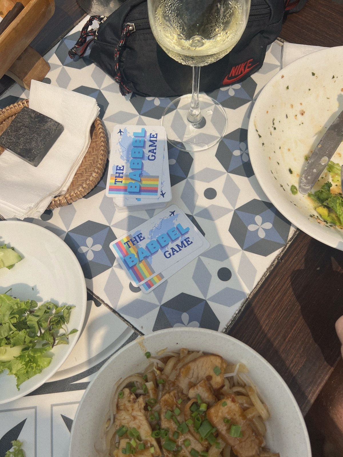Two Babbel Game boxes on a restaurant table between plates and a glass of wine