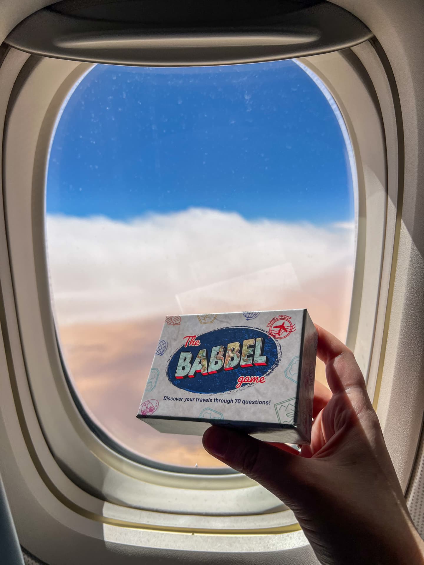 A hand holding The Babbel Game box in front of an aeroplane window above the clouds